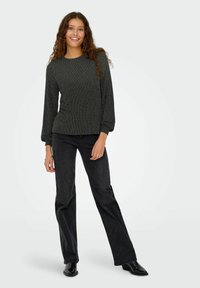 Zwart top met lange mouwen en een gestructureerd patroon, aansluitend bij de taille en uitlopend bij de manchetten. Gecombineerd met zwarte wijde broekspijpen en zwarte schoenen.