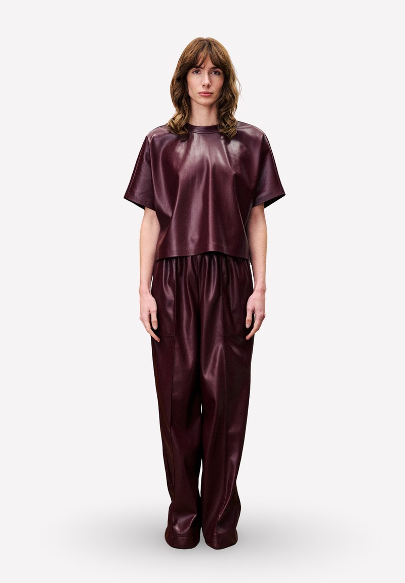 Vrouw staat rechtop, gekleed in een losse bordeauxrode leren top met korte mouwen en bijpassende brede broek tegen een effen achtergrond.