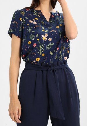 Femme portant un chemisier bleu marine à manches courtes à motif floral avec marguerites et feuilles, associé à un pantalon taille haute bleu marine noué avec une ceinture en tissu.