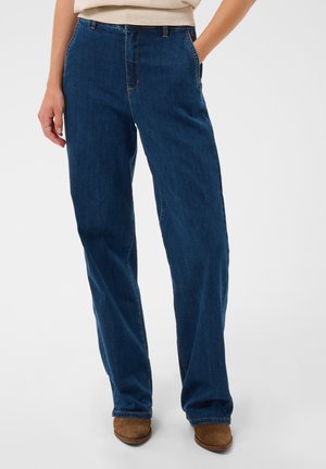 Femme portant un jean bleu foncé coupe droite, mains dans les poches et chaussures marron sur un fond uni