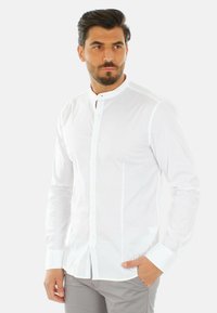 Camicia bianca a maniche lunghe con colletto a banda, dotata di una chiusura con bottoni e una texture morbida e liscia. Design slim fit con polsini sagomati.