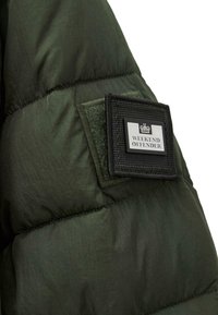 Chaqueta acolchada verde con paneles cosidos, que presenta un parche de tela negra etiquetado "WEEKEND OFFENDER" y un detalle de logo de corona.