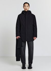 Cappotto nero imbottito con cappuccio, tasche frontali e taglio dritto, abbinato a pantaloni grigi e scarpe nere, tenuto insieme da una sciarpa nera.