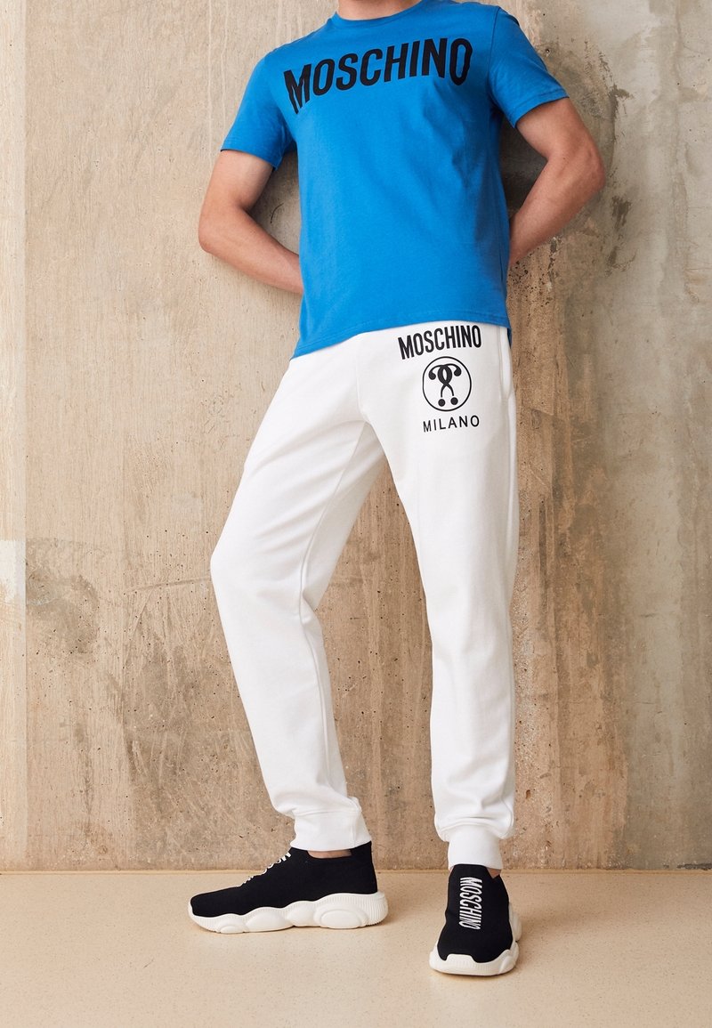 Muž oblečený v modrém tričku Moschino, bílých teplákách Moschino Milano a černých mokasínech Moschino proti texturované zdi.