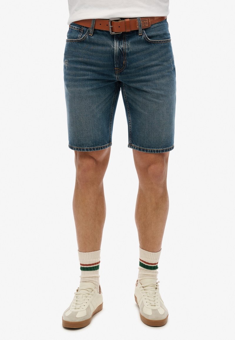 Shorts de mezclilla en azul oscuro con un corte regular, que presentan detalles de costura, cinco bolsillos y un acabado desgastado. Combinados con zapatillas de color claro.