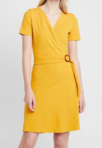 Gul omlottklänning med korta ärmar, V-ringning och en sköldpaddsaccent på midjebandet, tillverkad av mjukt, elastiskt tyg.