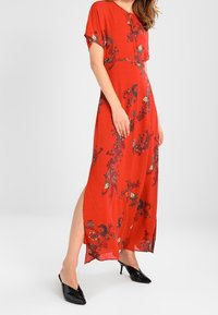 Rød maxikjole med blomsterprint, korte ærmer, sideskår og iført sorte spidse hæle.