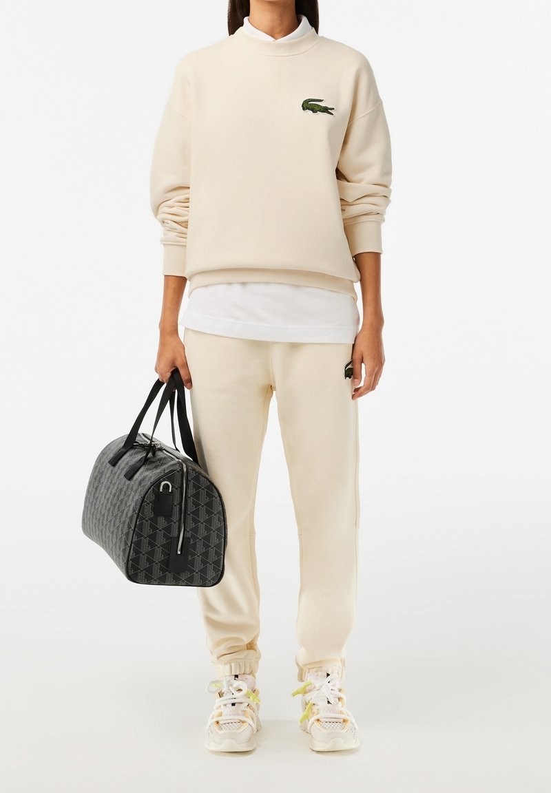 Personne portant un sweat-shirt beige Lacoste et un pantalon de survêtement, des baskets blanches, tenant un sac de sport gris foncé à motifs à côté d’elle.
