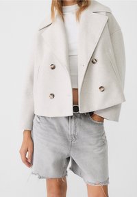 Light grey cropped jacket made of textured wool blend, featuring double-breasted design, large buttons, and notched lapels. Worn over denim shorts.