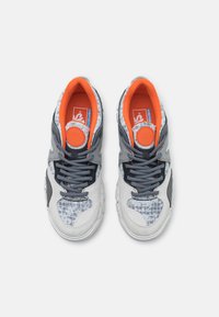 Baskets grises et blanches avec des accents orange, motifs texturés sur la boîte à orteils et les lacets. Semelle soutenante avec un détail de marque circulaire.