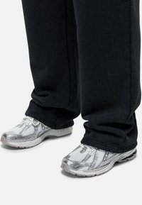 Zilveren sportieve schoenen met gaasdetails en een dikke witte zool, gedragen met losse zwarte sweatpants die gedeeltelijk de schoen bedekken.