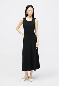 Γυναίκα φοράει ένα αμάνικο μαύρο μίντι φόρεμα με τετράγωνη λαιμόκοψη και λευκά μυτερά χαμηλότατα τακούνια, στέκεται μπροστά σε ένα απλό ανοιχτό φόντο.