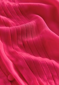 Tissu rose vif avec plis verticaux cousus et un petit bouton rond recouvert de tissu, présenté en plis doux et fluides.