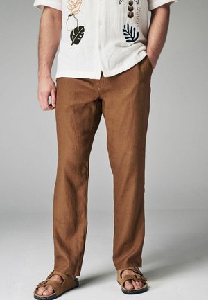 Pantaloni in lino marrone con una vestibilità ampia, abbinati a sandali scivolati marroni. L'outfit è caratterizzato da una camicia a maniche lunghe di colore chiaro con ricami.