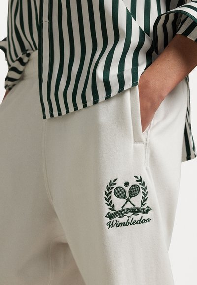 Valged dressipüksid külje taskutega, millel on vasakul reitel roheline brodeeritud Wimbledoni logo ja risti tennise reketid.