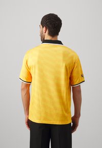 Gelbes Poloshirt mit gemusterter Textur, schwarzem Kragen und weißen Akzentstreifen an den Ärmeln. Mit Logo auf dem linken Ärmel.