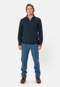 Pulôver de fleece azul marinho com colarinho alto e fecho de correr de um quarto; combinado com calças softshell azuis e botas castanhas com atacadores. Costuras planas visíveis.