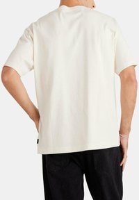 Camiseta de algodón de manga corta color crema, con un corte relajado, cuello redondo y un pequeño detalle de logo cerca del dobladillo. Vista trasera lisa.