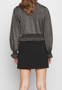 Kvinna som bär en långärmad topp i metallisk textur med rynkade manschetter och nederkant, matchad med en enkel svart A-linje kjol, vänd bort.