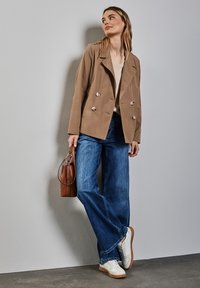 Trench coat doppiopetto beige con bottoni argentati, top beige chiaro, jeans in denim blu a gamba larga e sneakers bianche con dettagli grigi.