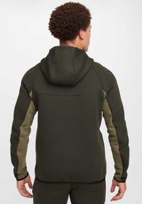 Khaki hoodie with a structured design, featuring a drawstring hood, contrasting olive-green panels on sleeves, and smooth textured fabric.