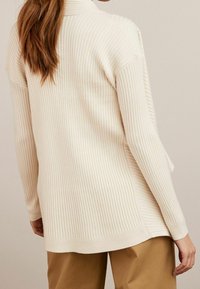 Pull long côtelé de couleur crème avec un col montant et des épaules tombantes, présentant un ourlet droit et des fentes latérales pour plus de confort.