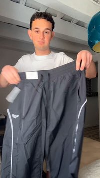 Jeune homme aux cheveux bouclés portant une chemise blanche tient un pantalon de sport noir avec des bandes réfléchissantes à l'intérieur.