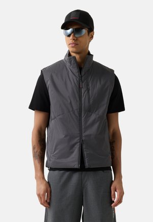 Man met zwarte pet, reflecterende zonnebril, zwart kort mouw shirt, grijze ritsvest en grijze broek met zichtbare tatoeages op zijn armen.