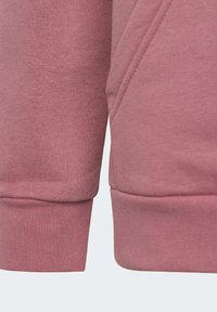 Detailní pohled na růžovou mikinu s texturovaným materiálem, žebrovanými manžetami a diagonálním švem. Navržena pro pohodlí a volnočasové nošení.