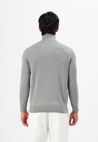 Mann in einem enganliegenden grauen Rollkragenpullover und weißen Hosen, der mit dem Rücken zu einer schlichten weißen Wand steht.