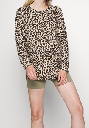 Kobieta ubrana w bluzkę z długim rękawem w panterkę i oliwkowo-zielone spodenki, stojąca z rękami przy krawędzi bluzki.