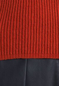Gros plan d'un pull tricoté rouge côtelé sur un pantalon en cuir noir lisse montrant le contraste des textures et les détails du tissu.