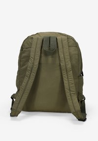 Mochila de nylon verde oliva con correas de hombro acolchadas, forma rectangular y un asa en la parte superior para colgar. Presenta costuras texturizadas y bolsillos laterales.