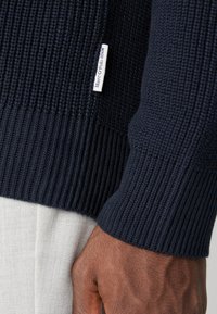 Marineblå strikkegenser med ribbestrikkede mansjetter og nederkant, med et lite hvitt merke med "Marc O'Polo DENIM" sydd på siden. Teksturert stoff.