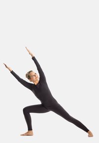 Black long-sleeve fitted yoga suit on a person, arms raised in a warrior pose, barefoot, against a plain white background.