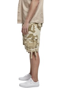 Cargo shorts i ett beige kamouflage-mönster, med flera sidofickor och remmar, kombinerade med vita sneakers.
