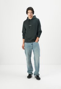 Donkergroene hoodie met een voorzak en een verstelbaar trekkoord, gecombineerd met lichtblauwe jeans en zwarte sneakers op een effen achtergrond.