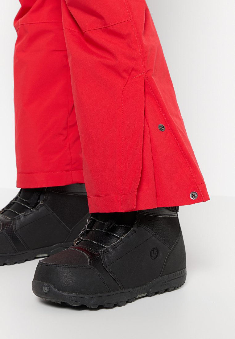 Stivali invernali neri indossati con pantaloni da neve rossi con chiusure a scatto alla caviglia su uno sfondo chiaro.