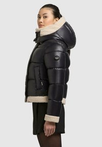 Puffige schwarze Jacke mit einem beigen Kunstfellkragen und -manschetten, seitlichen Reißverschluss Taschen und einem Logo-Patch am linken Ärmel. Leichtes Design.