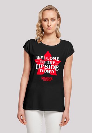 Schwarzes T-Shirt mit umgeschlagenen Ärmeln, das weißen und roten Text trägt, der "WILLKOMMEN IM GEGENTEIL" und "STRANGER THINGS" in fetter Schrift sagt.