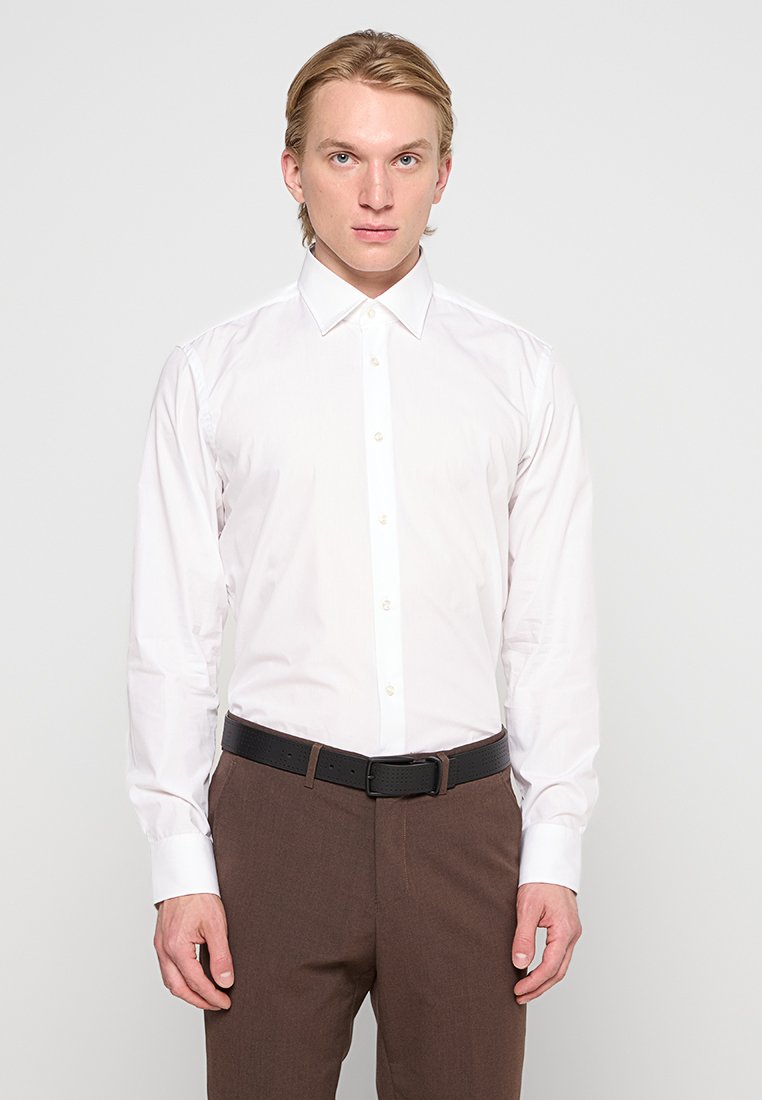 Chemise blanche à manches longues avec un col pointu, fermeture à boutons et coupe ajustée, portée avec un pantalon marron et une ceinture noire.