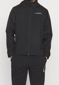 Musta Adidas Terrex -takki ja housut, joissa on täysi vetoketju ja kädet taskuissa.