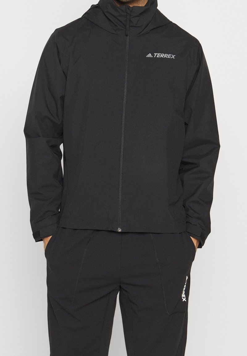 Musta Adidas Terrex -takki ja housut, joissa on täysi vetoketju ja kädet taskuissa.