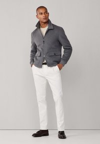 Mand iført en grå lynlåsjakke, cremet sweater, hvide bukser og mørkebrune læderloafers, stående mod en ensfarvet lys grå baggrund.