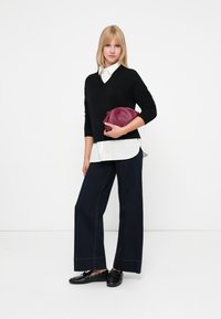 Suéter negro de pico, camisa blanca con cuello por debajo, jeans oscuros de pierna ancha y zapatos negros de slip-on. Sosteniendo un bolso burdeos.