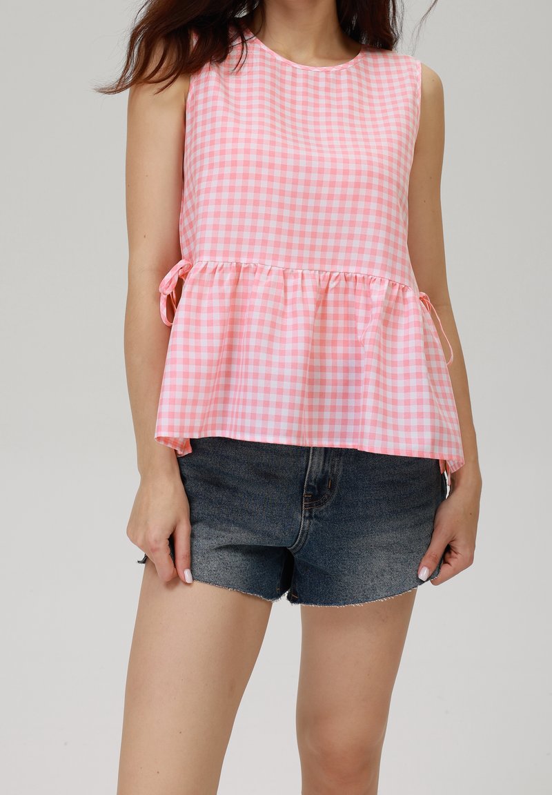 Femme portant un haut sans manches à carreaux rose et blanc avec des liens sur les côtés et un short en jean foncé sur un fond uni.