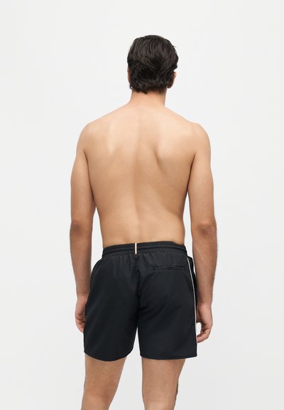 Homme debout de dos, torse nu, portant un short de bain noir avec une ceinture élastique et une poche arrière, sur un fond clair uni.