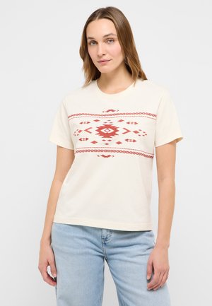 Kobieta o włosach do ramion, ubrana w kremową koszulkę z krótkim rękawem i czerwonymi geometrycznymi wzorami oraz jasnoniebieskie dżinsy, stojąca na jednolitym tle.