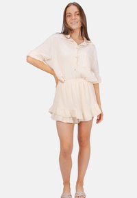 Light beige two-piece set featuring a button-up short-sleeve top with a pocket and a tiered ruffled skirt. Made of textured fabric.