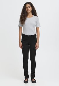 Junge Frau mit langen lockigen Haaren, die ein graues, kurzärmeliges, figurbetontes Oberteil, schwarze enge Jeans und schwarze Ballerinas trägt, vor weißem Hintergrund stehend.